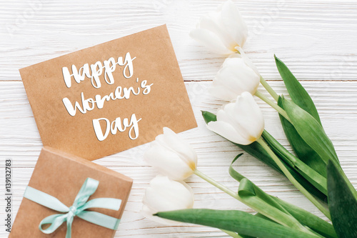 happy womens day text sign on stylish craft present with greeting card and tulips on white wooden rustic background. flat lay with flowers and gift with space for text. greeting card