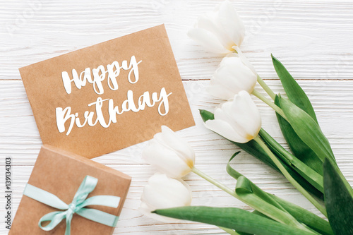 happy birthday text sign on stylish craft present with greeting card and tulips on white wooden rustic background. flat lay with flowers and gift with space for text. greeting card