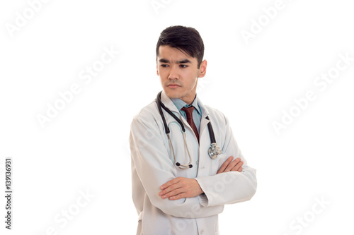 serious brunette male doctor in uniform with stethoscope on his neck posing with crossed hands isolated on white background