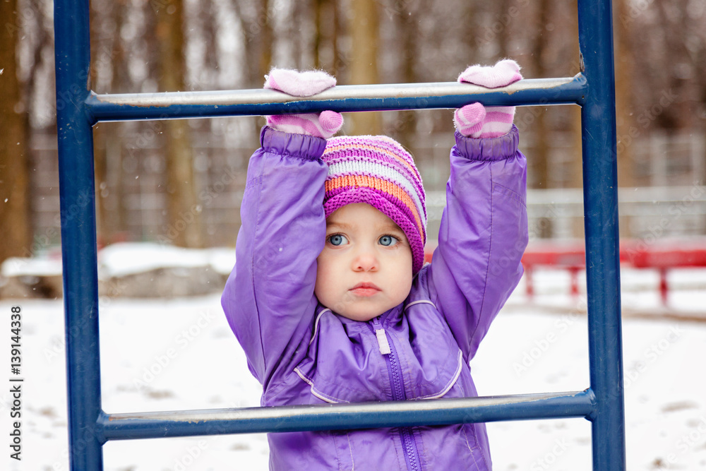 Fototapeta premium Portrait of little girl one year old playing outside in winter park playground. Toddler girl looking straight to the camera. Snowy weather.