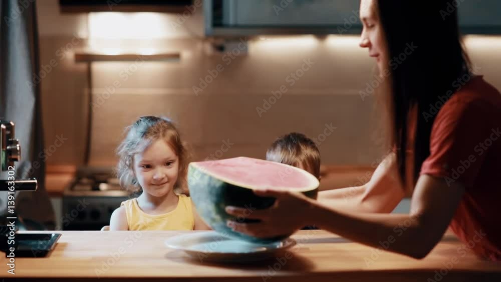 Brother and sister sitting at the table, young mother brings a half of watermelon and put it on the table. Brother and sister sitting at the table, young mother brings a half of watermelon and put it on the table.