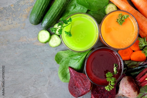 Three glasses of healthy vegetable juice with scattered ingredients, above view over a slate background