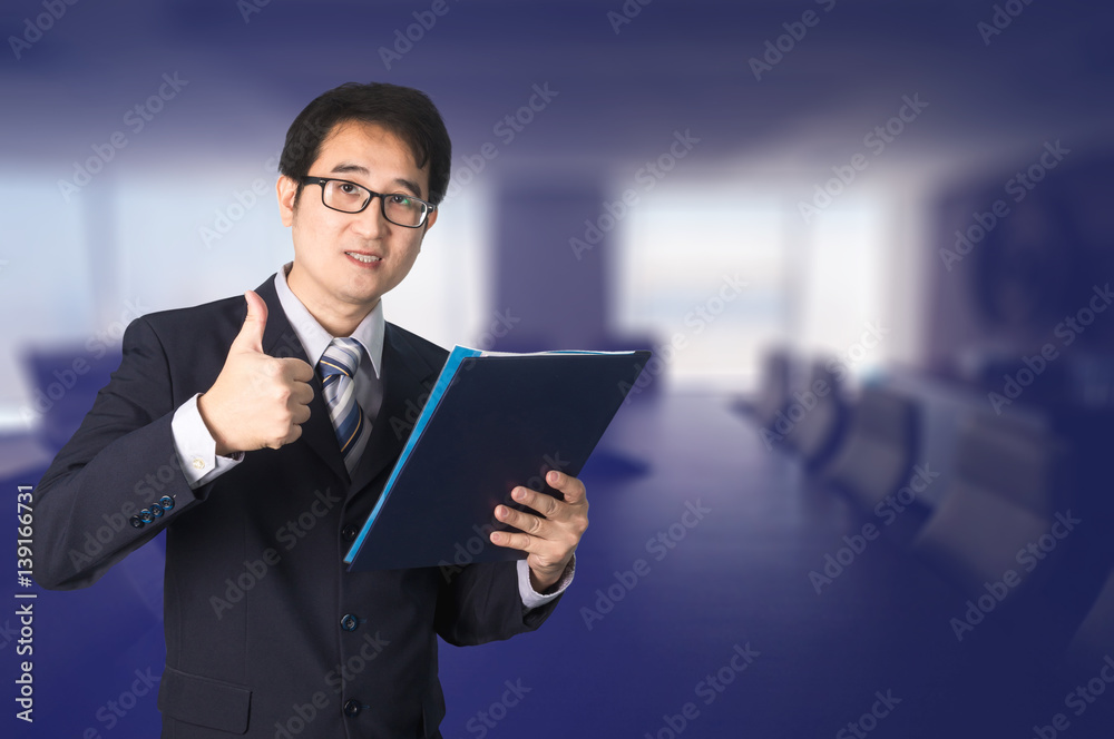 Confident Asian businessman showing document file and giving thumbs up in front of meeting room.