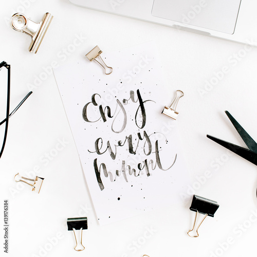 White office desk workspace with quote Enjoy Every Moment and office supplies. Laptop, scissors, pen, clips, glasses on white background. Flat lay, top view.