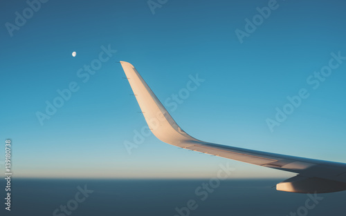 View from illuminator of dark teal morning misty horizon  clear sky during dawn without clouds  wing of modern airplane and partly closed moon