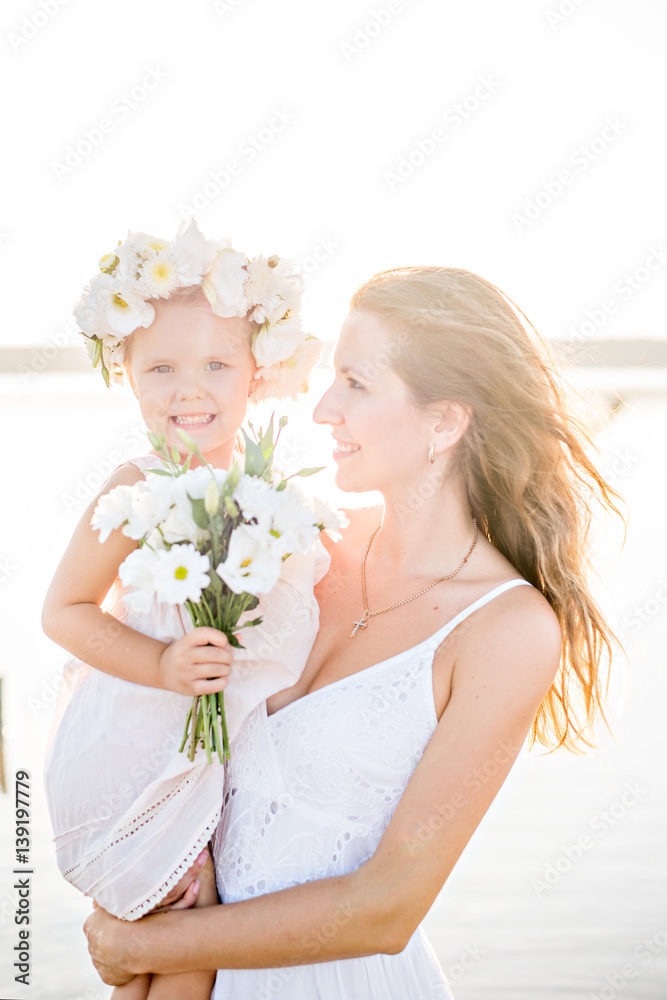 Mom and daughter in white dresses with garlands of flowers on her head at sunset Mom and daughter in white dresses with garlands of flowers on her head at sunset