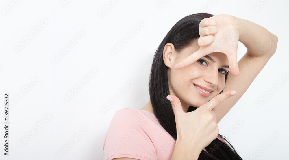 Look here. Attractive young woman showing frame hand and looking at the camera to see the world. Portrait of happy girl your hands up on white.