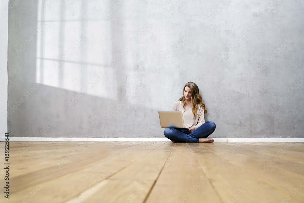 Blond woman sitting on the floor in front of grey wall using laptop Blond woman sitting on the floor in front of grey wall using laptop