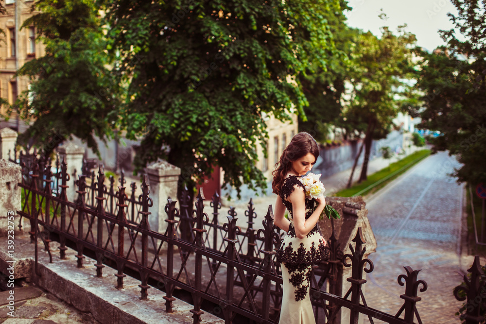 Naklejka premium Girl in fancy evening dress holds bouquet of peonies posing on bridge with steel handrails