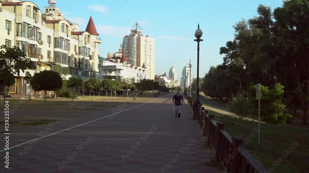 A young man is jogging ouside in the city across the path. The male is wearing trendy leggins and shorts outfit. He runs near the buildings along the path. A young man is jogging ouside in the city across the path. The male is wearing trendy leggins and shorts outfit. He runs near the buildings along the path.