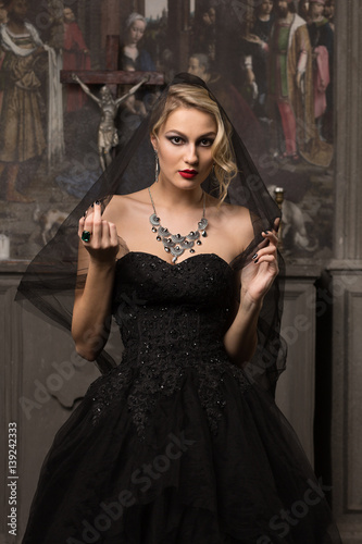 beautiful blonde woman in a black evening dress stands on a background of the altar and paintings on his head wearing a black veil around her neck jewelry, looking at the camera