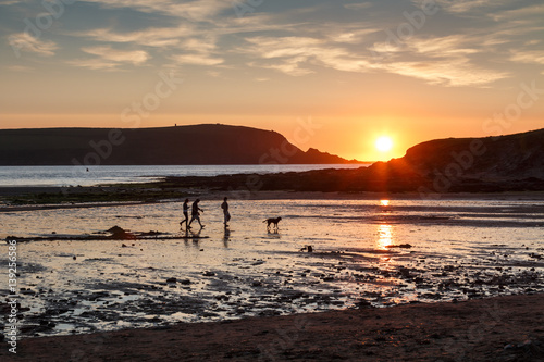 A family with dog in silhouette walking on a beach at Daymer Bay in Cornwall England.  The setting sun is reflecting on the wet sand and the sea in the Camel estuary.