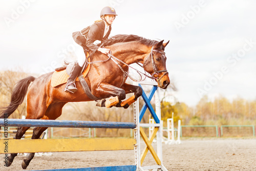 Obraz na plátně Bay horse with rider girl jump over hurdle on show jumping competition
