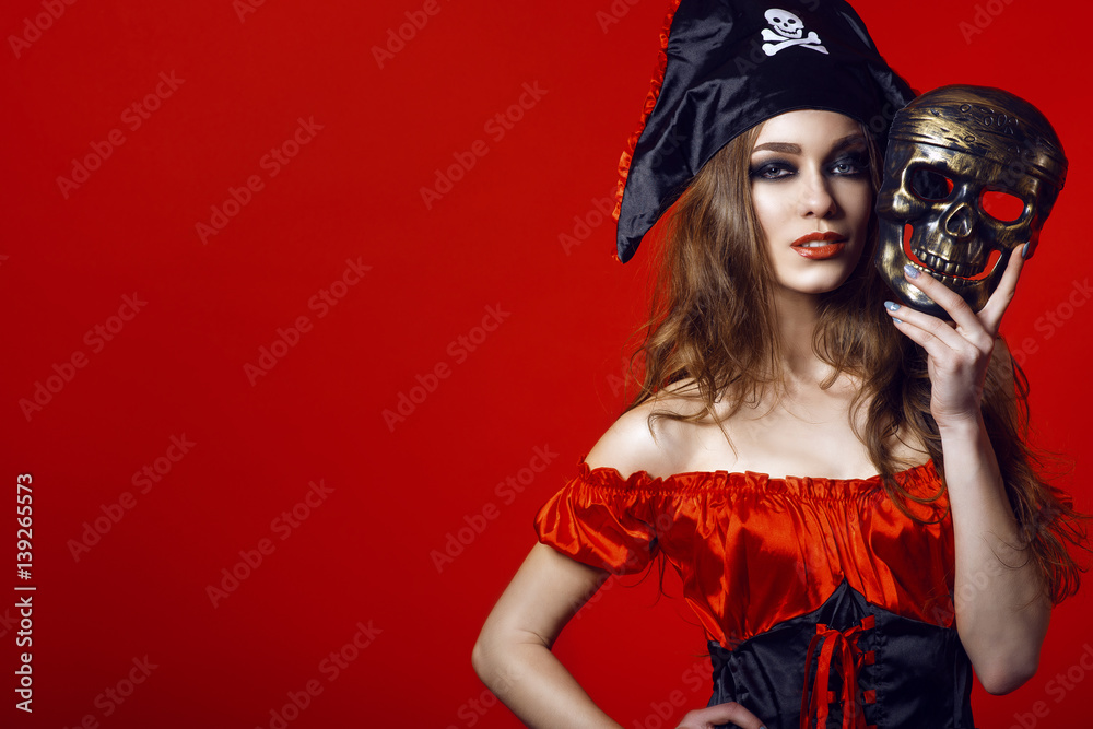 Naklejka premium Portret wspaniała seksowna kobieta z prowokującym makijażem w pirata kostiumu mienia czaszki masce obok jej twarzy. Koncepcja hazardu i kasyna. Pojedynczo na jasnym czerwonym tle. Ścieśniać. Kopiowanie przestrzeni