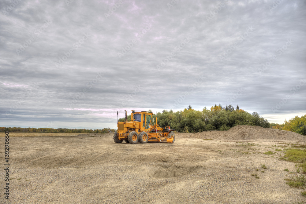 Fototapeta premium An orange industrial gravel road grader parked at a small gravel pit by an autumn forest in a rural countryside landscape