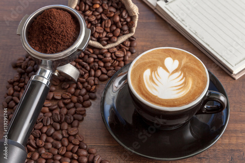 Obraz na płótnie Coffee cup of latte art in the black color cup with some coffee beans, portafilter and notebook on wooden background w salonie