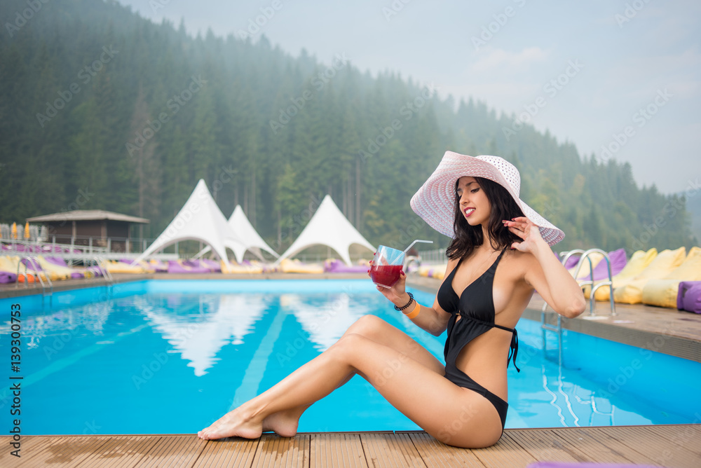 Young tender woman in a hat and black bikini sitting next to swimming pool and drinking cocktail on the background of mighty forest