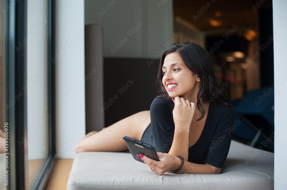 Smiling girl with perfect body in black bodysuit lying on mattress near big window and holding in hands tablet computer on vacation at lounge zone in the spa resort Smiling girl with perfect body in black bodysuit lying on mattress near big window and holding in hands tablet computer on vacation at lounge zone in the spa resort
