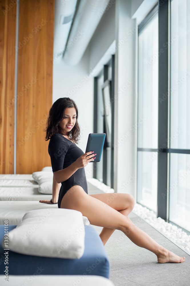 Attractive beautiful female wearing black bodysuit sitting on white lounger near big windows and smiling looks at tablet computer at spa resort Attractive beautiful female wearing black bodysuit sitting on white lounger near big windows and smiling looks at tablet computer at spa resort