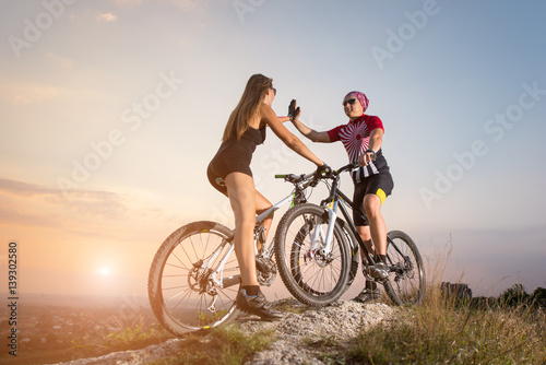Couple bikers gives a high five to each other against the backdrop of a magnificent sunset. Cyclists standing with the mountain bicycles on the rock
