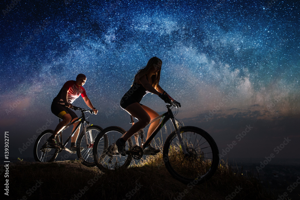 Obraz premium Woman and man riding a bicycles on the hill in night. Starry sky with Milky way over the two mountain bikers. Bottom view