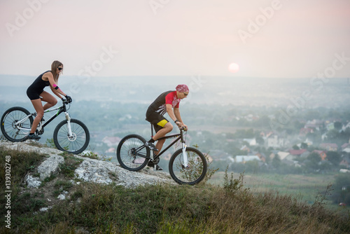 Cyclist with girlfriend in motion on sports bicycles on the background of beautiful sunset. Pink Kinesio tape glued on the girl's hand. Below is a city in the distance. Blurring background