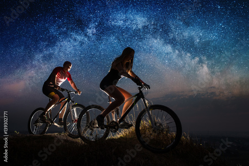 Woman and man riding a bicycles on the hill in night. Starry sky with Milky way over the two mountain bikers. Bottom view