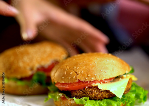 Hamburger fast food with ham on wooden board . Group of hamburger on blurred background. Piece of cheese hanging from sandwich. Human hand reaches for cheeseburger is not in field.