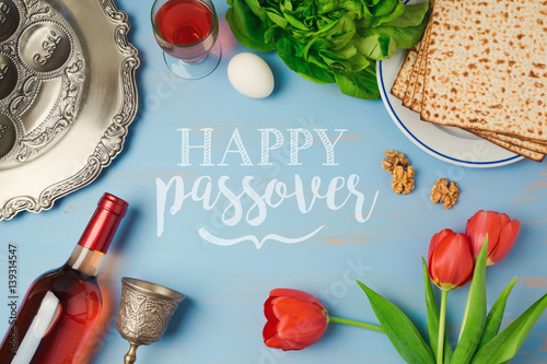 Passover holiday greeting card with seder plate, matzoh, tulip flowers and wine bottle on wooden background. Top view from above