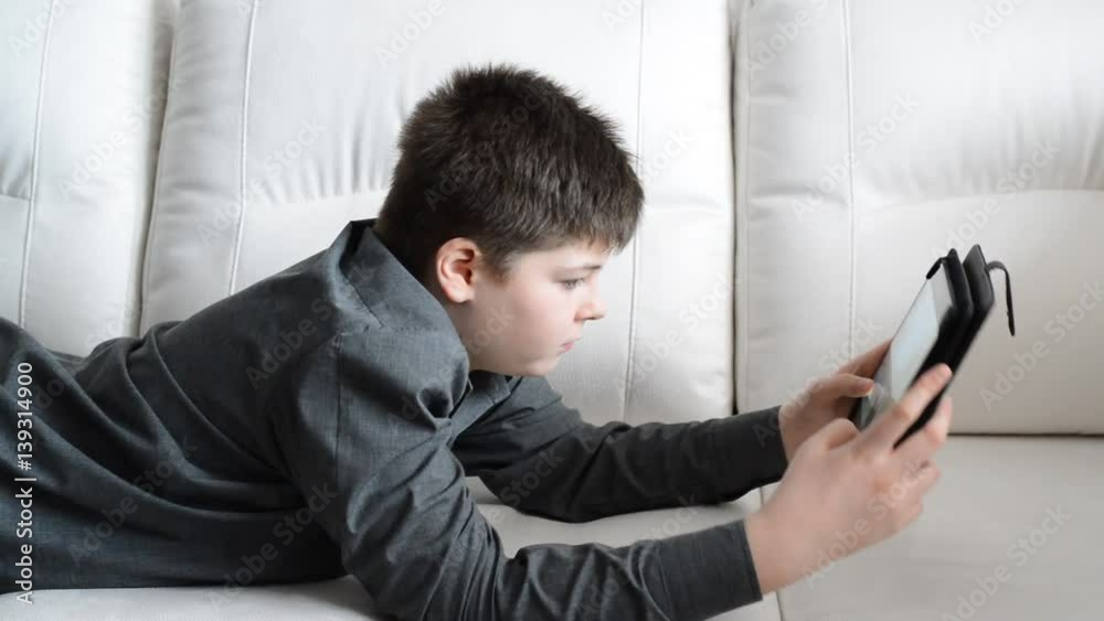 The teenager playing on tablet lying on a sofa in room The teenager playing on tablet lying on a sofa in room