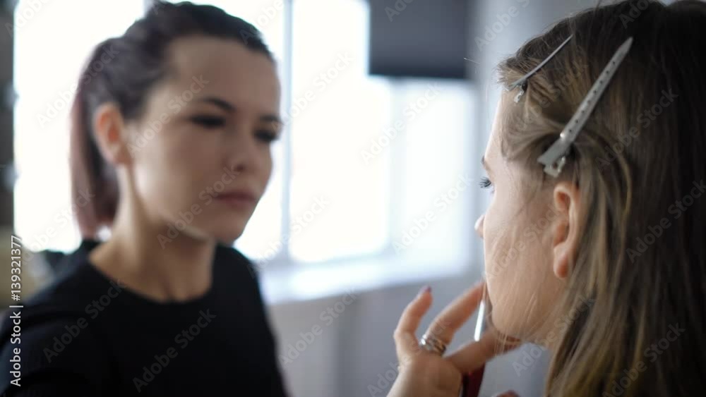 Professional make-up artist in the fashion industry is doing makeup young model who is sitting on a chair in her beauty studio. Girl with a brush in his hands blurred on the background.