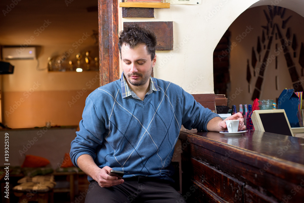 Young man is having coffee in bar, sitting and playing with his phone