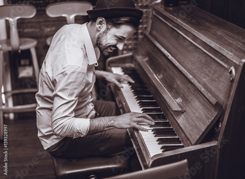 Fototapeta Trendy man with stylish hat and beard trying playing vintage old wooden piano