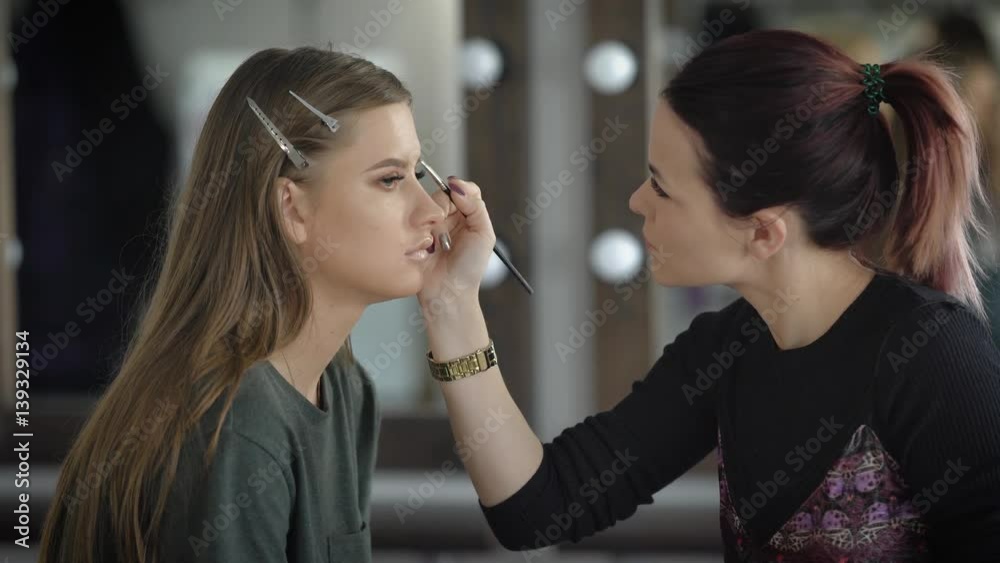Close up of make-up process at beauty shop. Young visagiste is applying eyeshadow on model's face using brush. Woman is sitting on the chair and brunette artist is creating an image of model. Close up of make-up process at beauty shop. Young visagiste is applying eyeshadow on model's face using brush. Woman is sitting on the chair and brunette artist is creating an image of model.