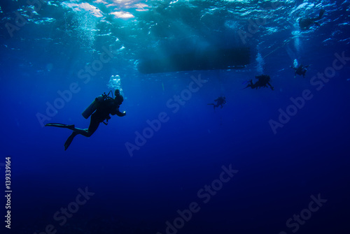 Scuba divers hang out underneath a boat are lit by the sun's light beams that break through the clear blue water.  The wavey surface can be seen above the people and bubbles are released © drew