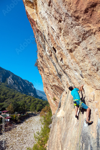 Mature Climber hanging on Rock above Forest and Village vertical