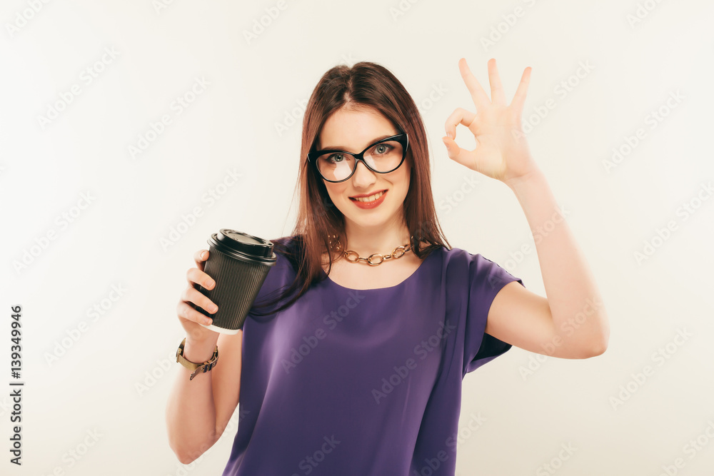 young beautiful girl in glasses with cup of coffee from a fast food smiling on a gray background. hand gestures. young beautiful girl in glasses with cup of coffee from a fast food smiling on a gray background. hand gestures.