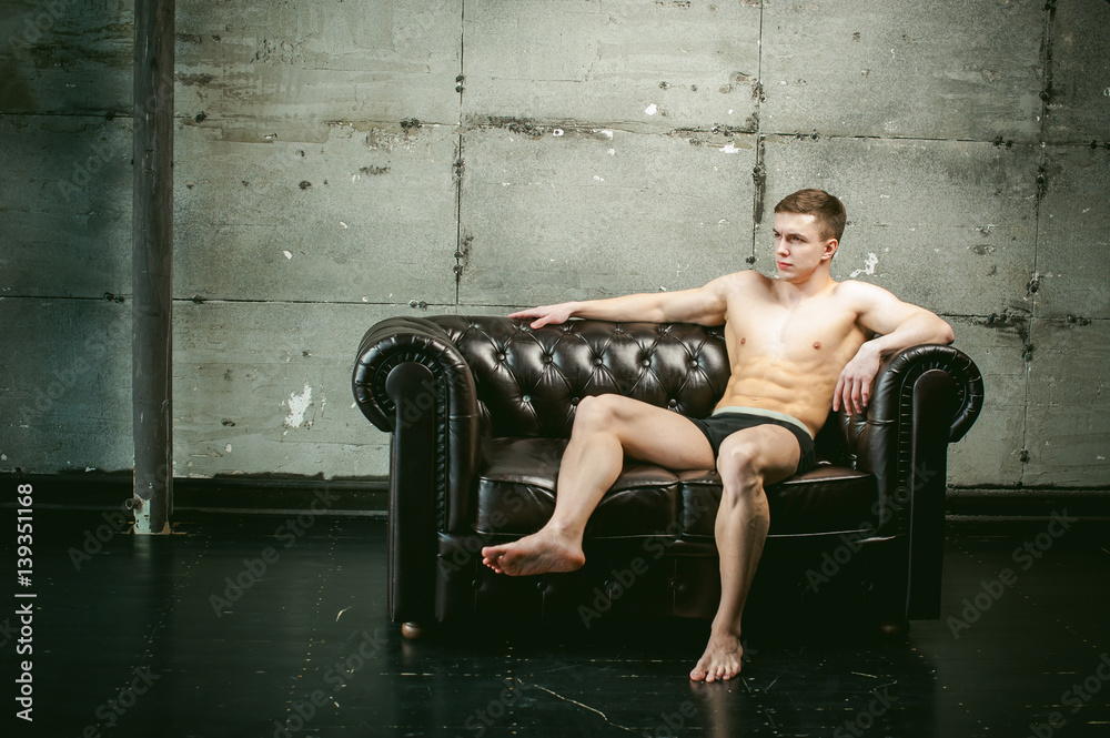 Young sexy men bodybuilder athlete sitting on a black leather couch with a naked torso, studio portrait