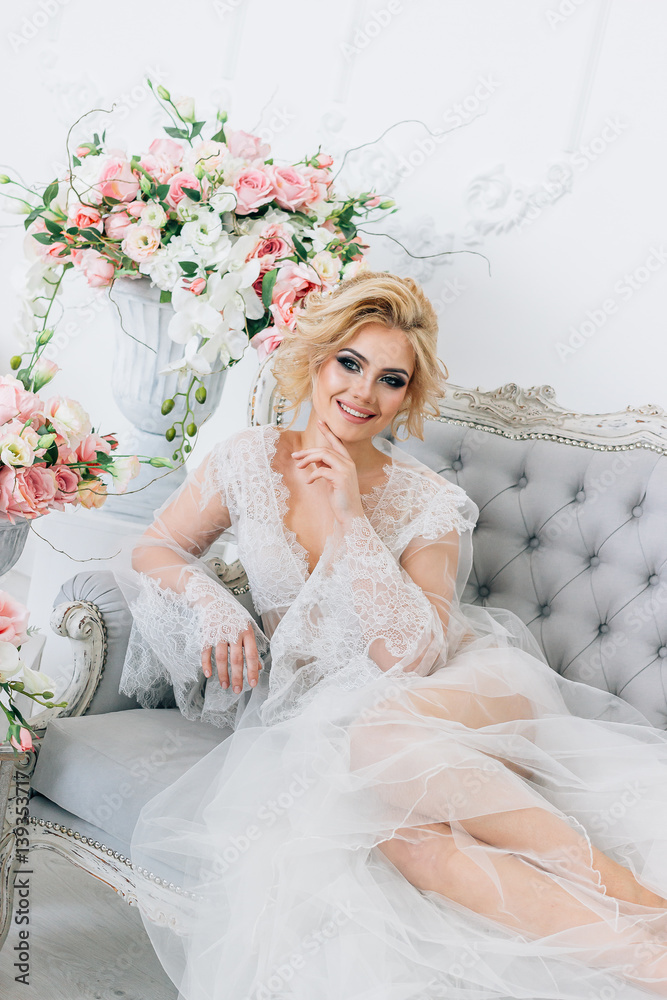 Beautiful girl with blond hair in a peignoir elegantly sits on the couch surrounded by flowers Beautiful girl with blond hair in a peignoir elegantly sits on the couch surrounded by flowers