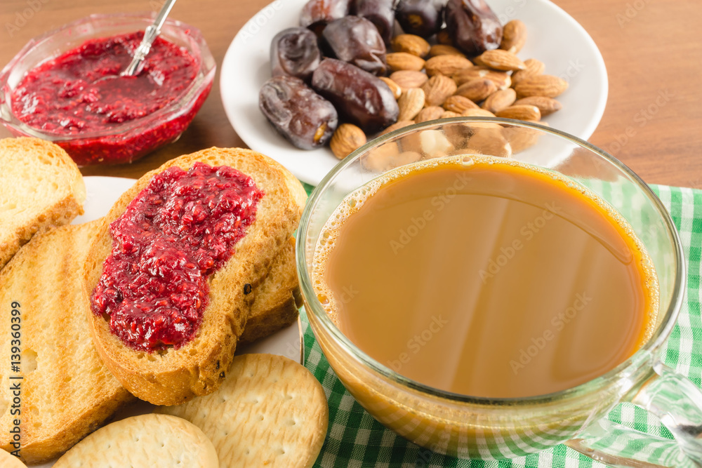 Breakfast time, cup of coffee with milk, toasts, raspberry jam, dates, almonds at napkin on wooden table. Breakfast time, cup of coffee with milk, toasts, raspberry jam, dates, almonds at napkin on wooden table.