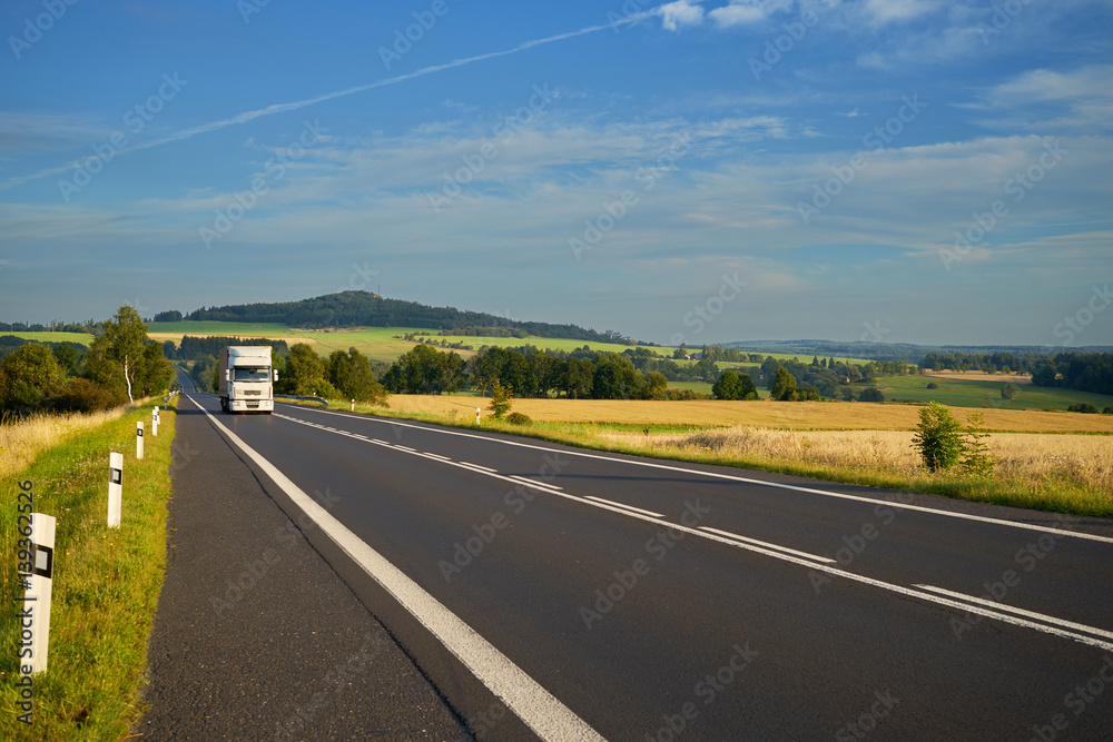 Fototapeta premium White truck arriving from a distance on the asphalt road between agricultural fields in a rural landscape