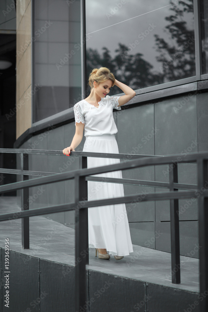 Fototapeta premium Blond hair girl in white dress stands on the metal stairs, on the gray background wall. Smiling and posing in front of a window.