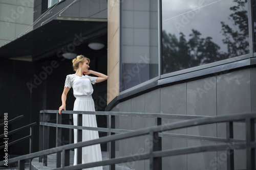 Blond hair girl in white dress stands on the metal stairs, on the gray background wall. Smiling and posing in front of a window.