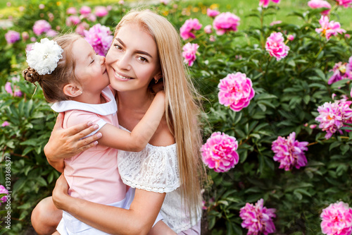 Happy mother and daughter in peony garden. Child kissing mother. Happy family outdoors. Trinity. Pentecost. Mother's day