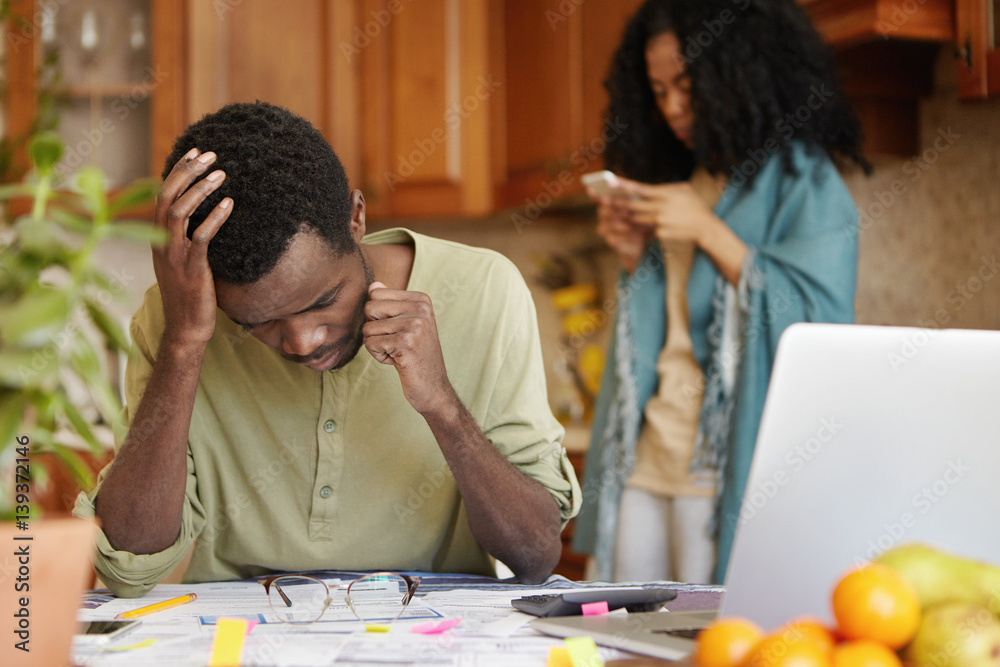 Unemployed young African-American man facing financial stress, feeling depressed and frustrated, holding head in despair, not able to pay off debts, his wife standing in background using mobile
