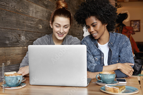 Happy cheerful interracial lesbian couple enjoying online communication on generic laptop computer, having video call to their friends during lunch at modern coffee shop, looking and smiling at screen