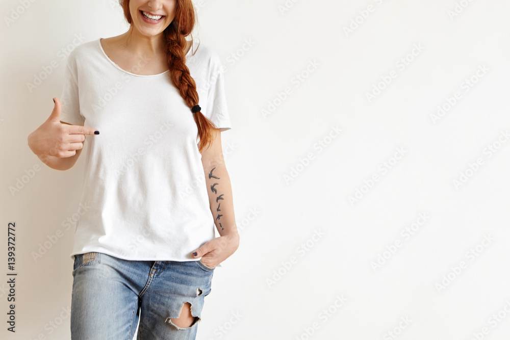 Fashion, clothing and design concept. Cropped portrait of attractive redhead European teenager with tatoo, smiling cheerfully, pointing finger at copy space on her stylish white oversize t-shirt Fashion, clothing and design concept. Cropped portrait of attractive redhead European teenager with tatoo, smiling cheerfully, pointing finger at copy space on her stylish white oversize t-shirt