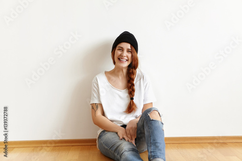 Indoor shot of beautiful happy Caucasian student girl with charming smile wearing white t-shirt, hat and ragged jeans, sitting on floor, leaning on blank wall while relaxing at home after university © wayhome.studio 