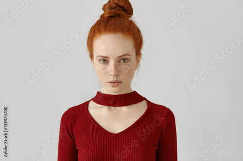 Beauty, style, fashion, clothing and design concept. Gorgeous luxurious Caucasian redhead woman with hair bun and freckles wearing stylish red dress with geometrical cut out before going out on date