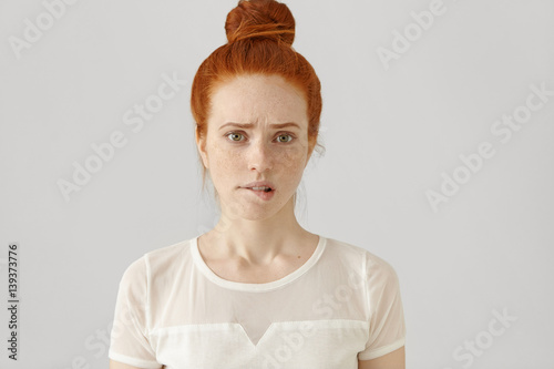 Confused or puzzled beautiful young Caucasian woman with ginger hair frowning, biting her lower lip after having done something wrong, looking at camera with guilty and apologetic facial expression photo